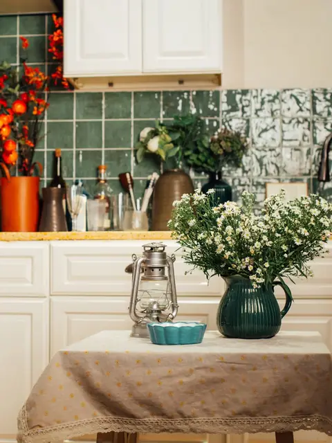 A small kitchen table with a vase of white flowers in front of green tiles and white cabinets