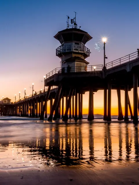 Huntington Beach Pier and its lifeguard tower at sunset, reflected on the wet sand