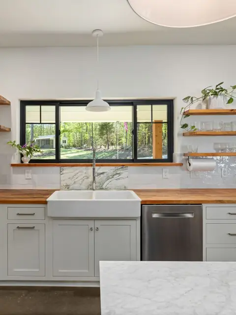 A bright kitchen with open wood shelves, a farmhouse sink and a black framed window onto trees